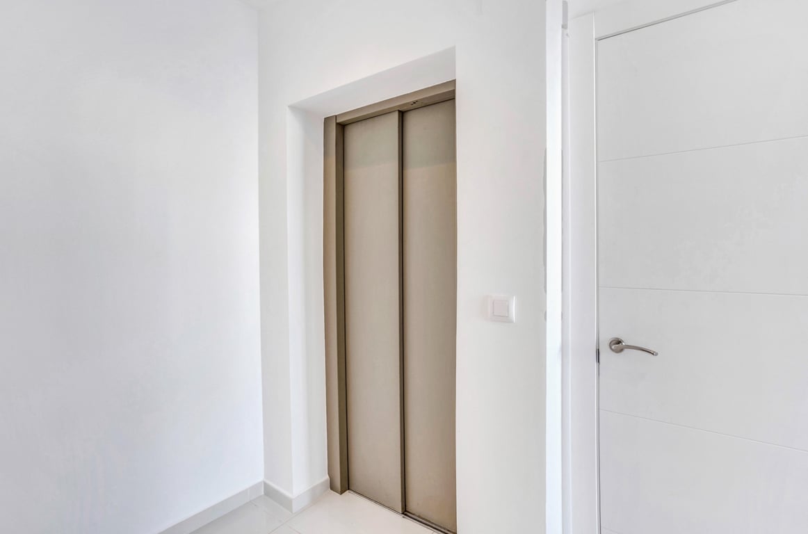 The private elevator with bronze-toned metallic doors is integrated into a minimalist hallway featuring white walls and matching interior woodwork.