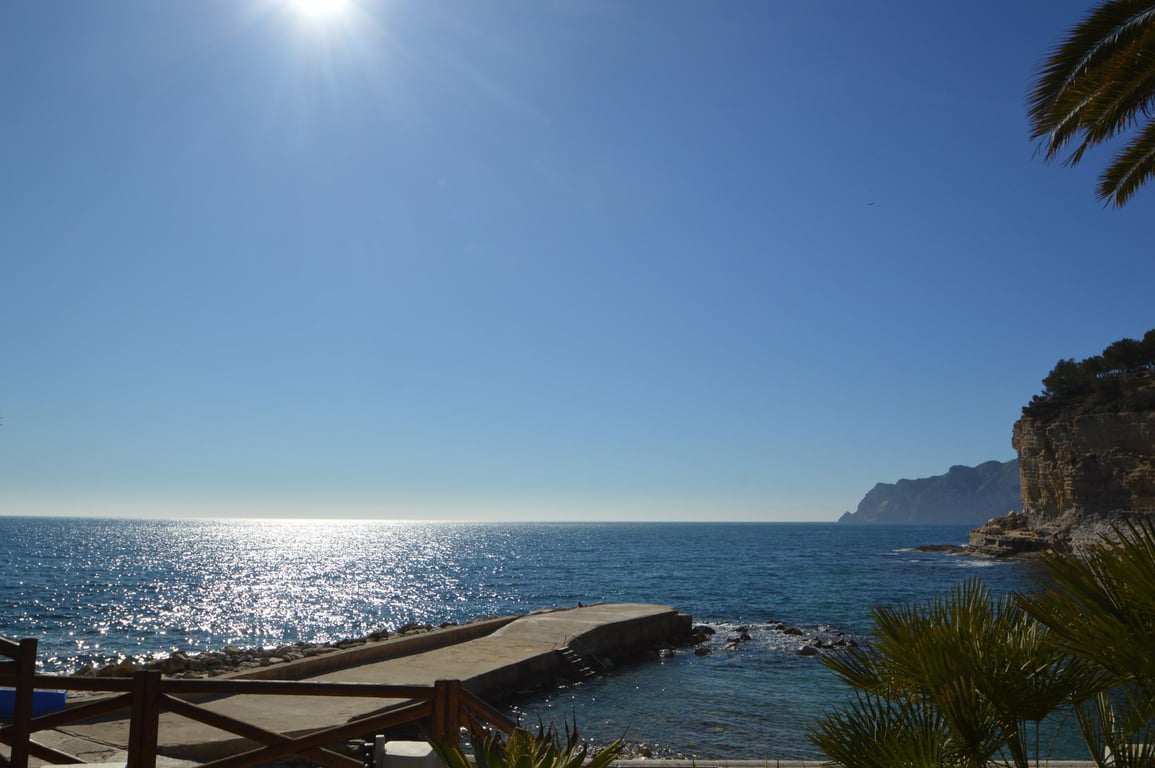 View of Cala Advocat featuring a concrete jetty and sparkling Mediterranean waters. The scene includes rocky cliffs and native palm trees under a bright sun.