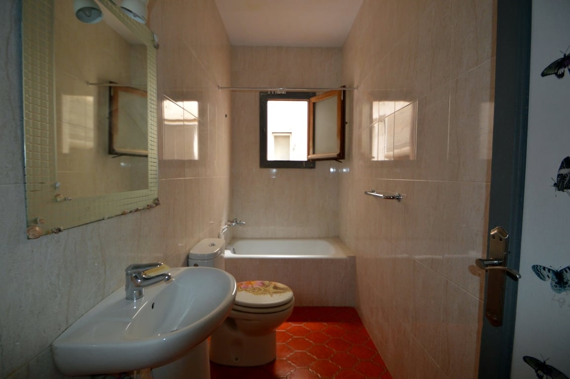 Bathroom featuring red hexagonal floor tiles, cream wall tiling, a pedestal sink, and a built-in bathtub positioned under a small window for natural ventilation.