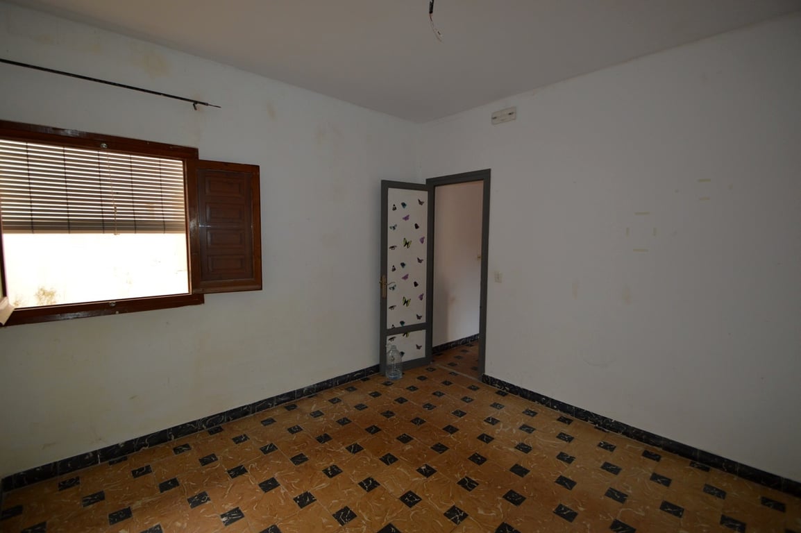 Bedroom featuring traditional patterned floor tiles, white walls, and a wooden window with shutters. A versatile space ready for renovation.