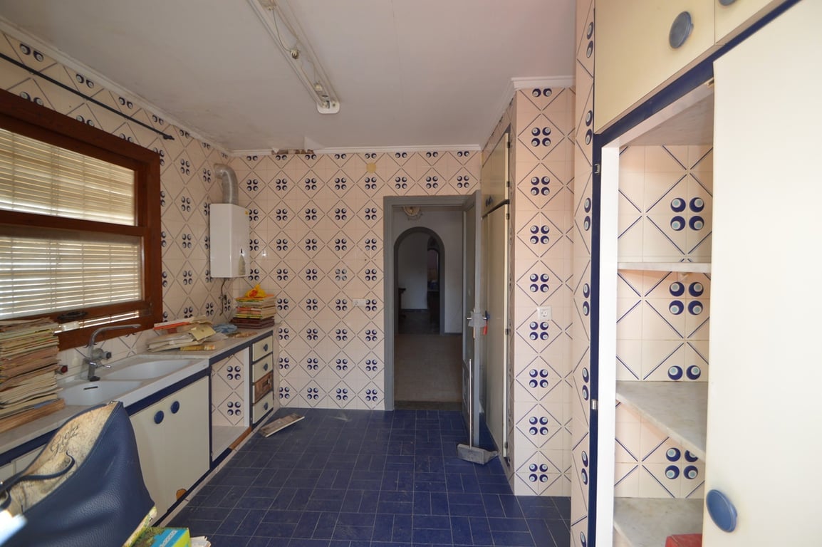 Kitchen featuring patterned wall tiles, blue floor tiling, and white cabinetry with blue accents. Includes a double sink under a wooden window and access to an arched hallway.