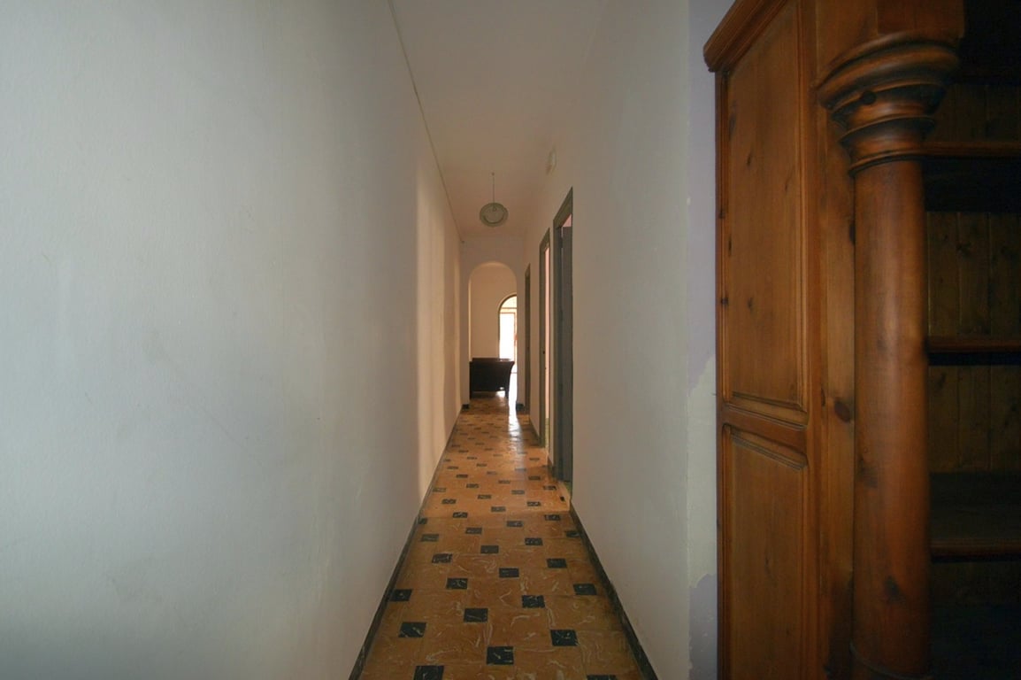 Long hallway featuring traditional patterned tile flooring and white walls, leading towards an arched doorway and a solid wood cabinet with carved details.