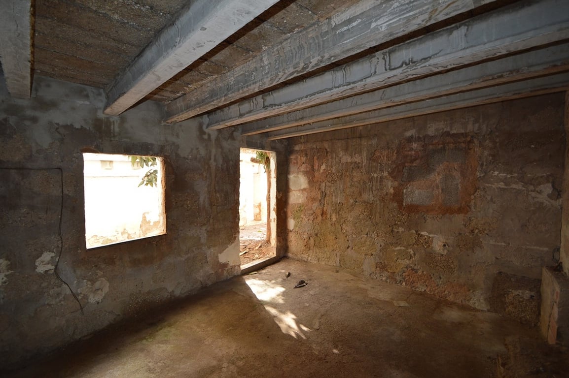 Raw interior space featuring original stone walls and exposed ceiling beams. Natural light enters through a window opening and a doorway leading to the courtyard.