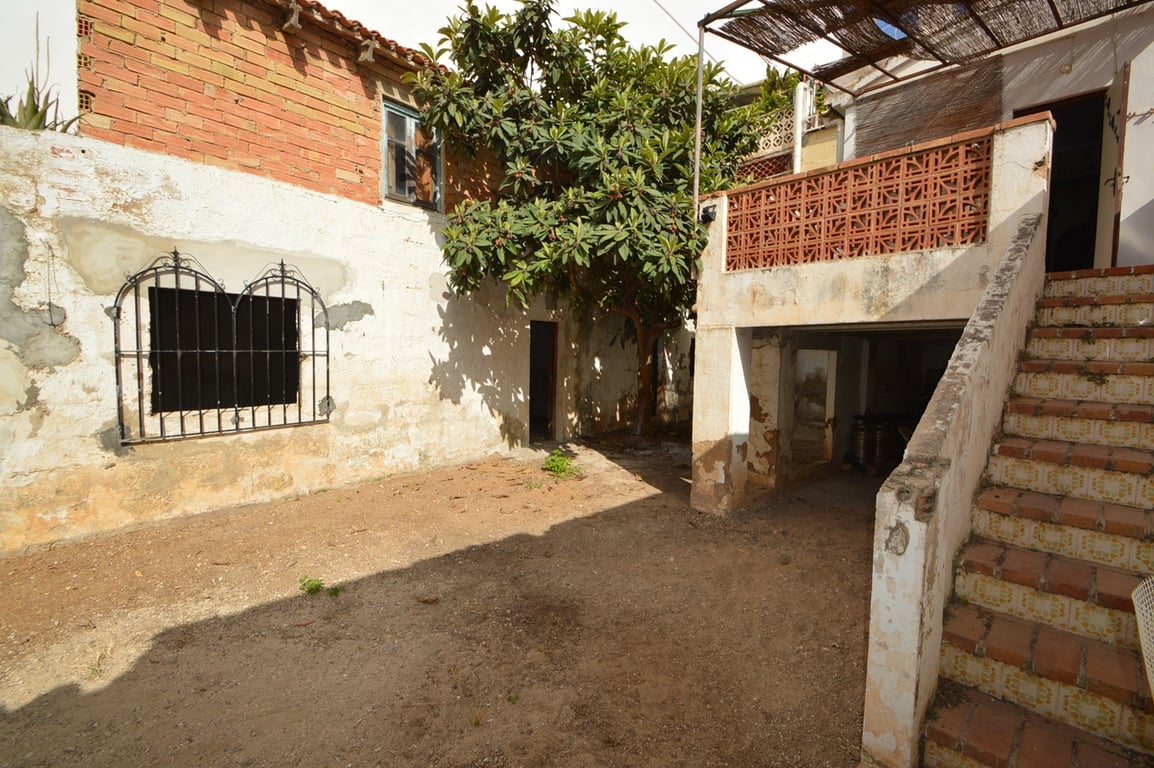 Rustic inner courtyard featuring a dirt floor, a mature tree, and outdoor tiled stairs leading to a terrace with a reed canopy and ceramic railings.
