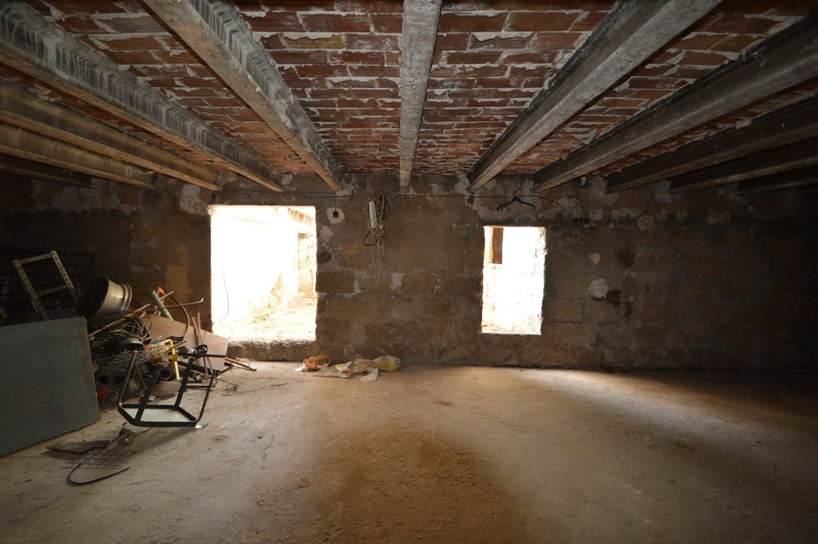Spacious open-plan interior for renovation featuring concrete beams and exposed brick ceilings. The room includes natural stone walls and two openings providing natural light.