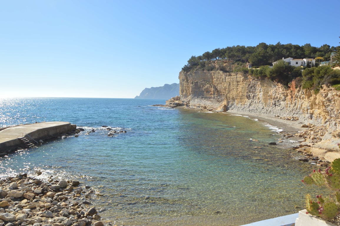 Scenic view of Cala Advocat featuring transparent Mediterranean waters, rugged limestone cliffs with pine trees, and a small concrete pier in Benissa.