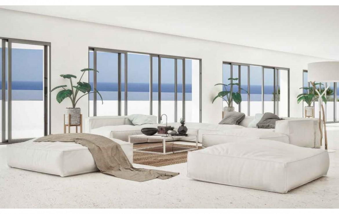 Minimalist living area featuring white modular floor seating, light stone flooring, and expansive sliding glass doors overlooking the Mediterranean horizon.