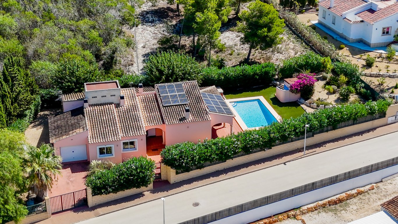 Aerial view showing the villa's tiled roof with solar panels, the private pool area with artificial turf, and the gated entrance with garage on a flat, enclosed plot.
