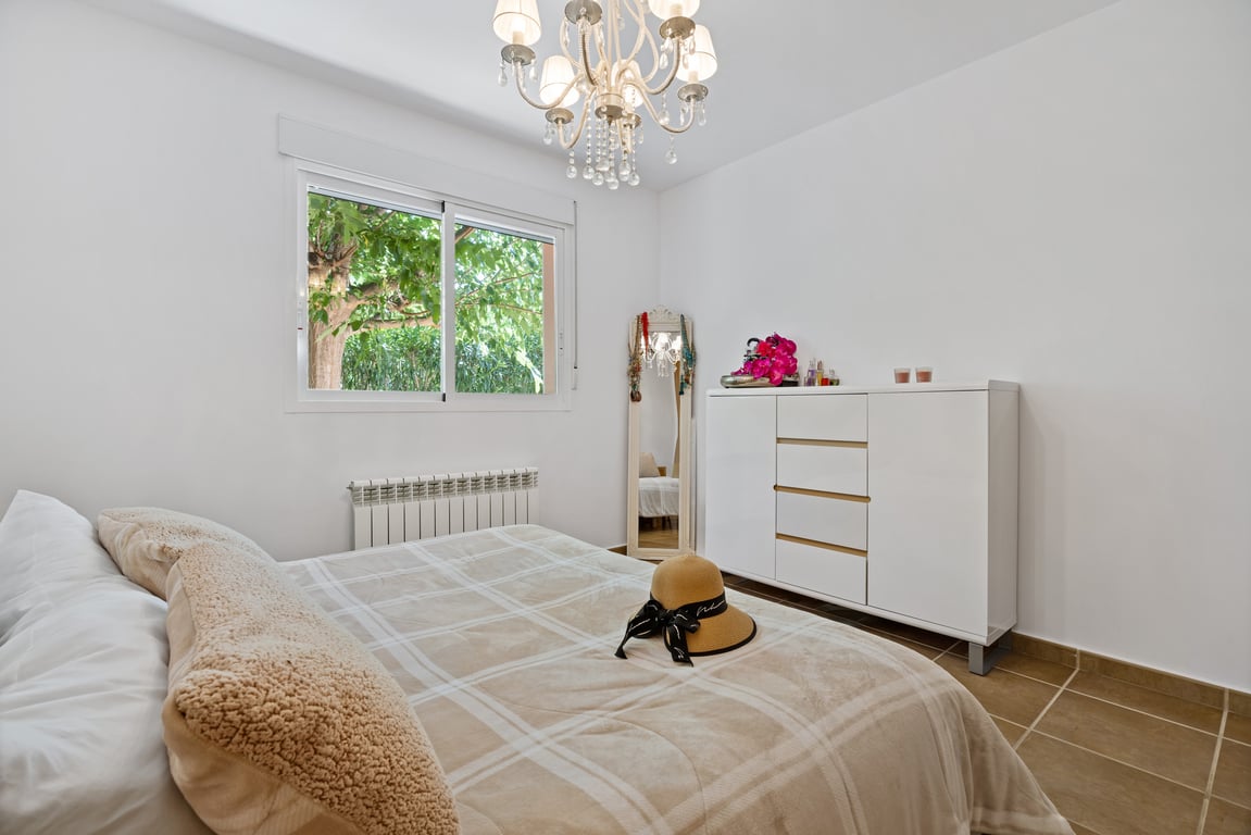 Bright bedroom featuring tiled flooring, a double-glazed window overlooking greenery, central heating radiator, and an ornate crystal chandelier.