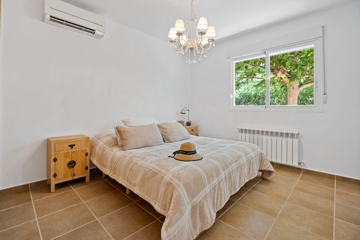 Bright bedroom featuring tiled flooring, split-system air conditioning, and a radiator. A large window offers views of the surrounding greenery and natural light.