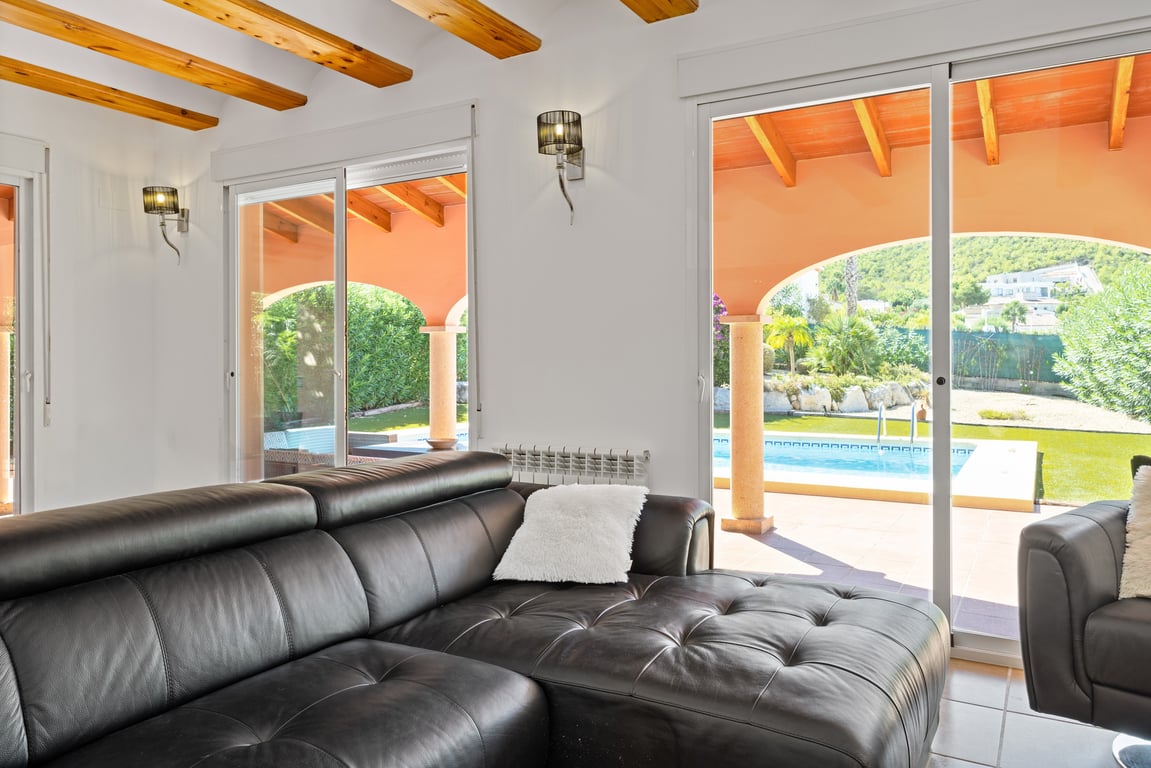 Bright living area featuring exposed wooden ceiling beams, a black leather sectional, and sliding glass doors opening to the pool and arched terrace.