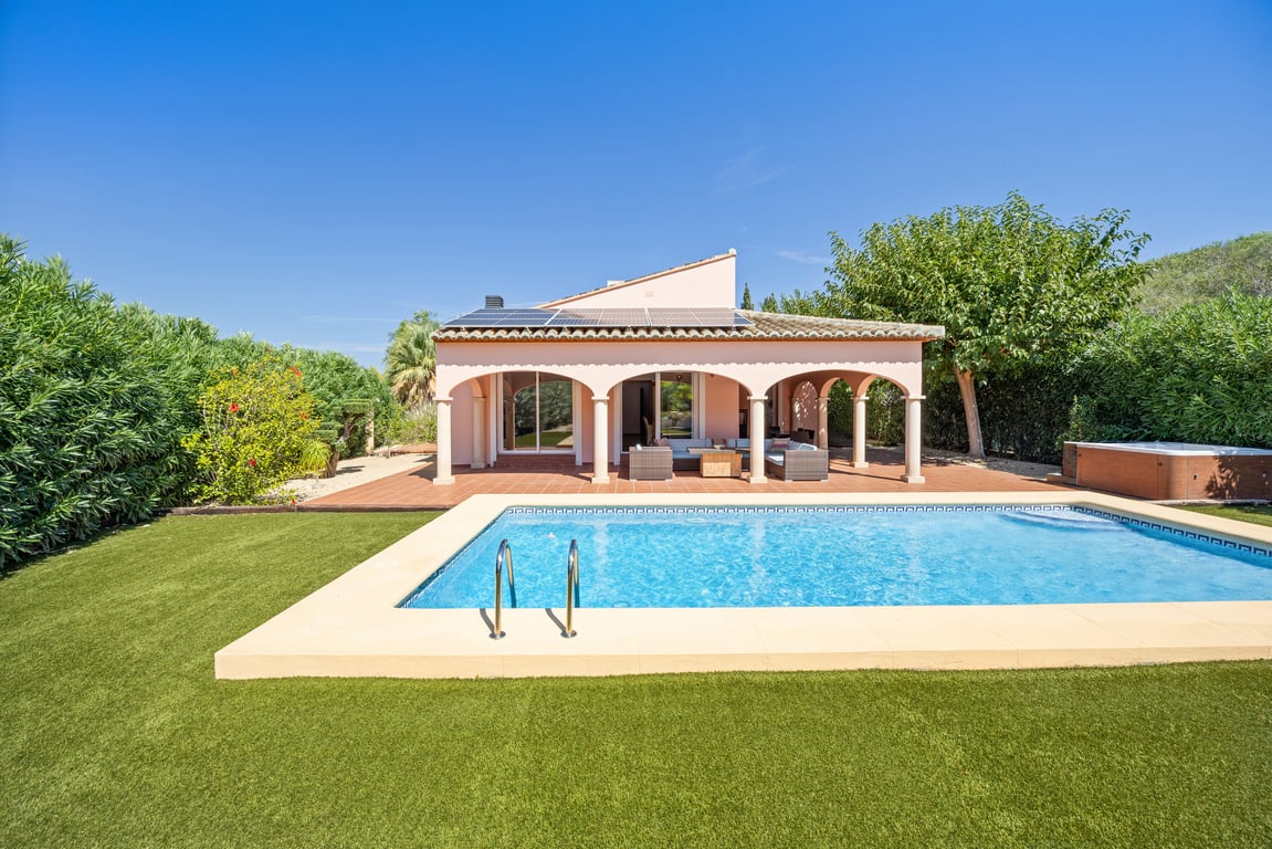 Exterior view of the villa featuring a mosaic pool, artificial lawn, and arched naya. Notable features include roof solar panels and a side jacuzzi.