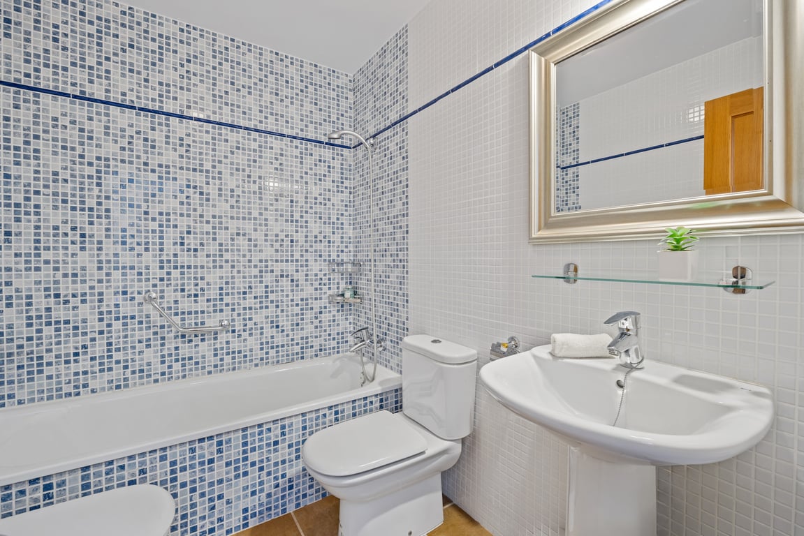 Functional bathroom featuring a bathtub with blue and white mosaic tiling, a white pedestal sink, and a large silver-framed mirror.