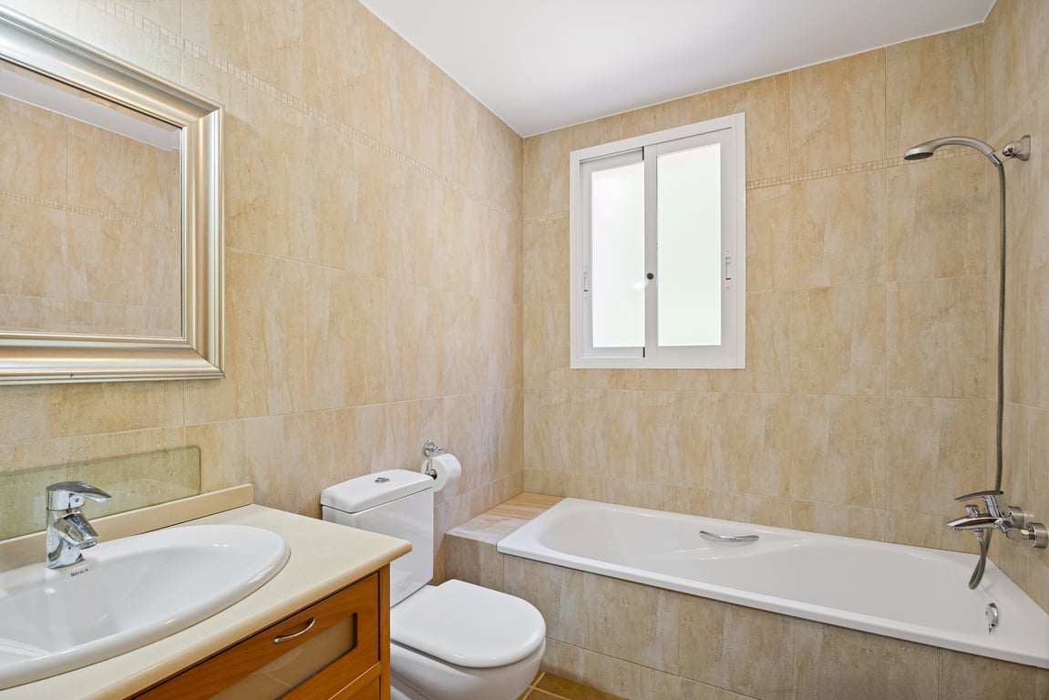 Functional bathroom featuring beige wall tiles, a built-in bathtub with shower, a wooden vanity unit, and a frosted window for privacy and natural light.