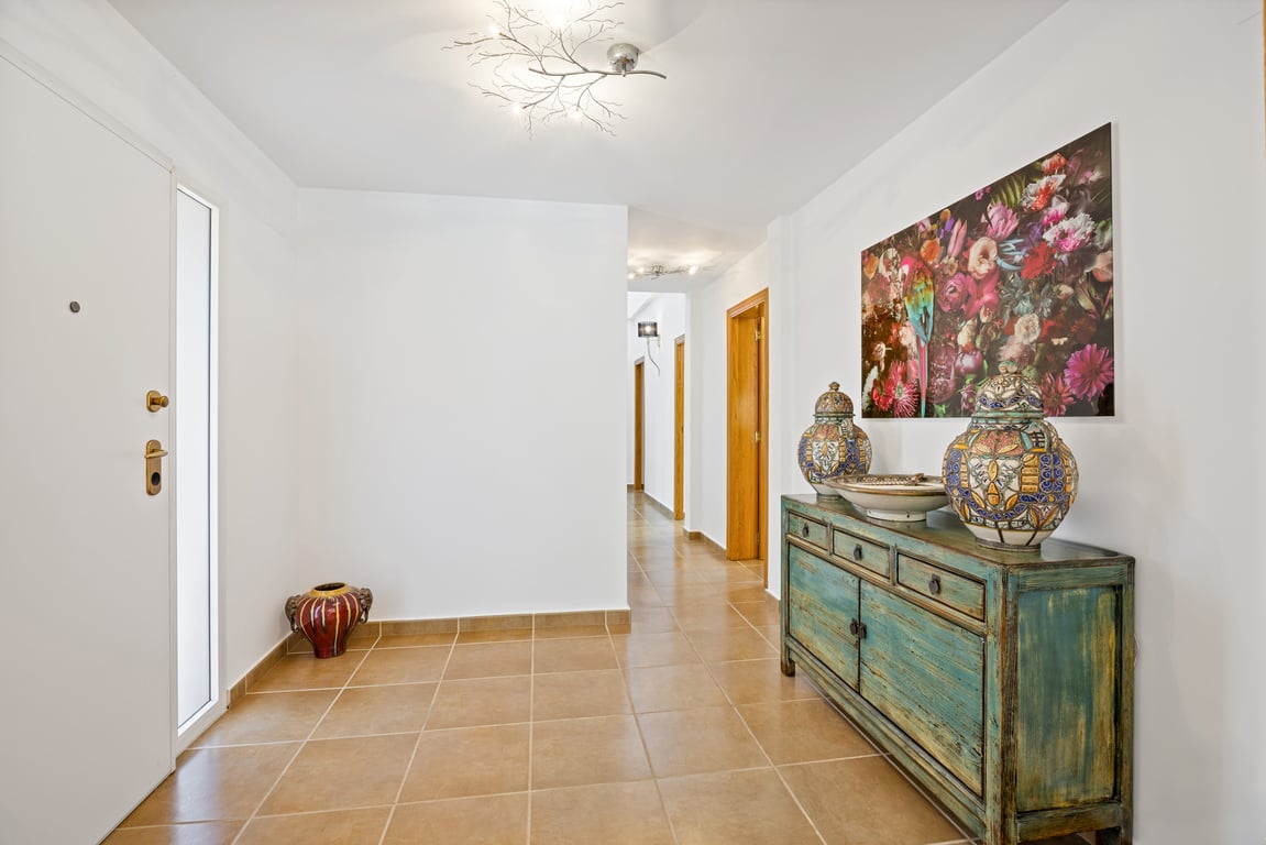 Spacious entrance hall featuring terracotta-style floor tiles, a turquoise wooden sideboard, and a bright corridor leading to the bedrooms with contemporary branch-style ceiling lights.