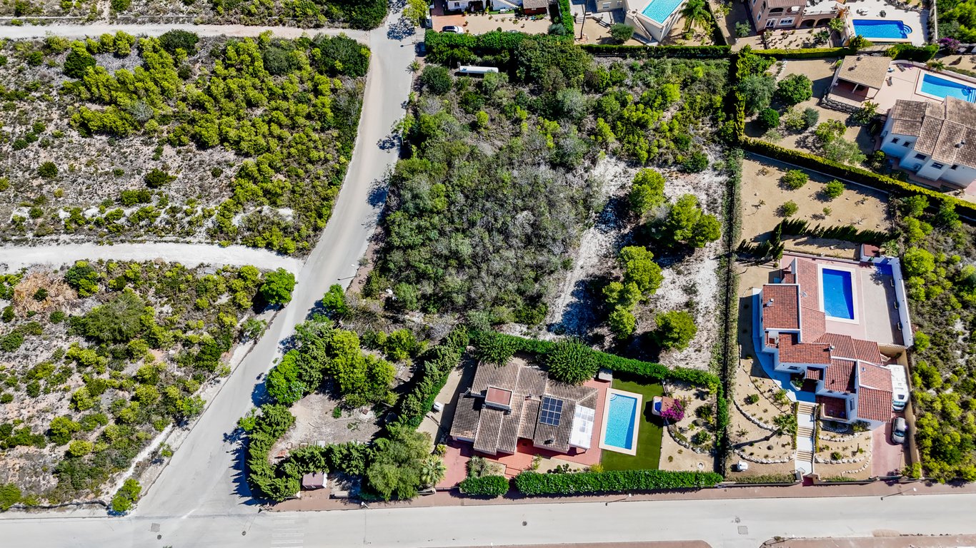 Aerial perspective of the villa featuring solar panels and pool, alongside the large adjacent urban plot with natural vegetation in Jávea.