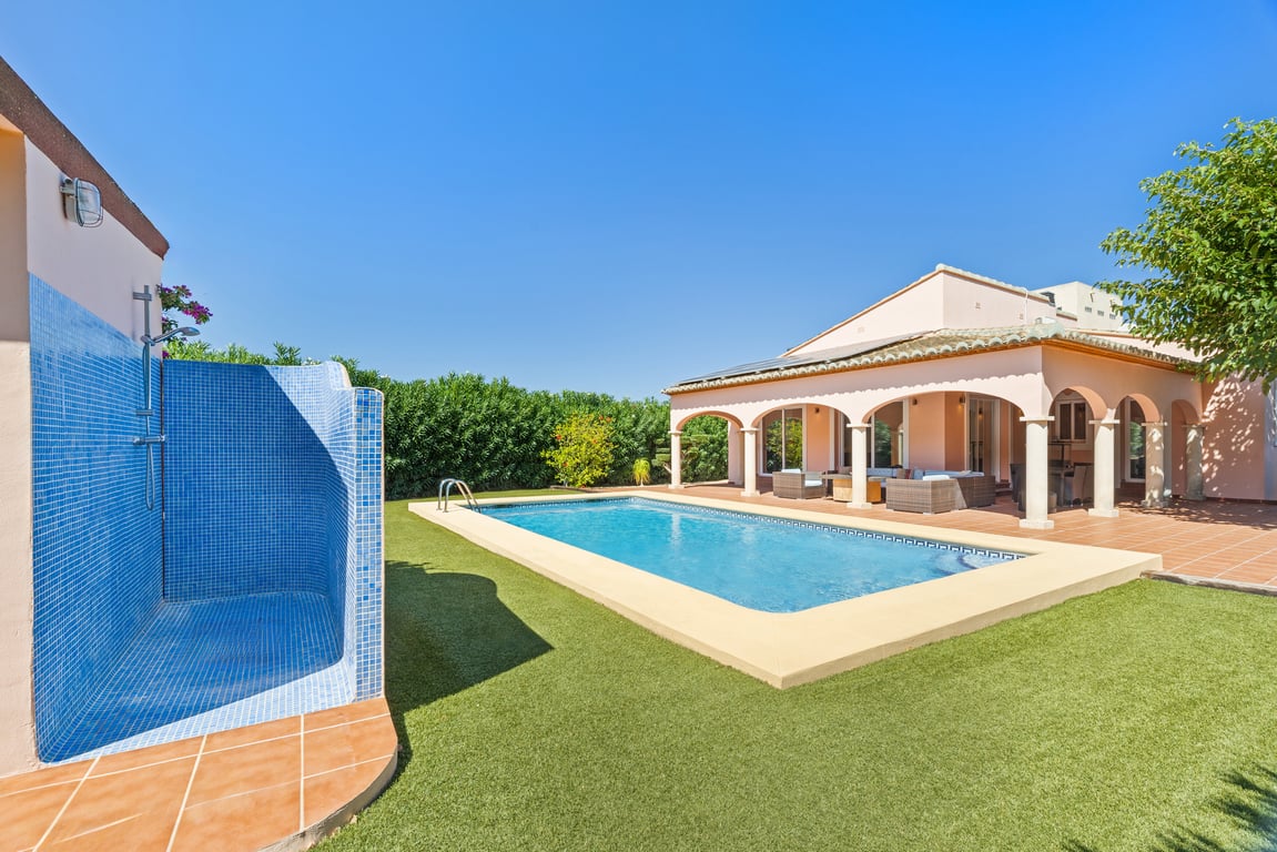 Blue mosaic swimming pool bordered by artificial turf, featuring an outdoor shower with tile cladding and a spacious arched naya with comfortable seating.