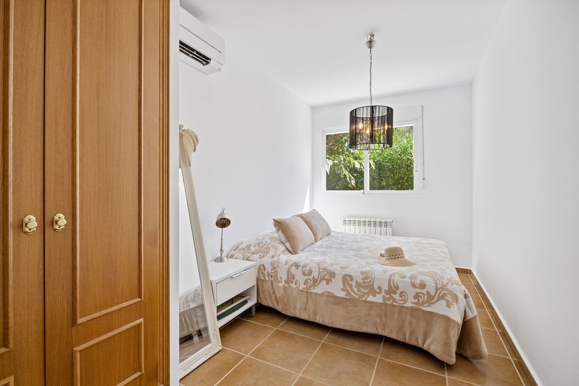 Bright bedroom featuring terracotta tile flooring, split air conditioning, a built-in wooden wardrobe, and a window overlooking the greenery.