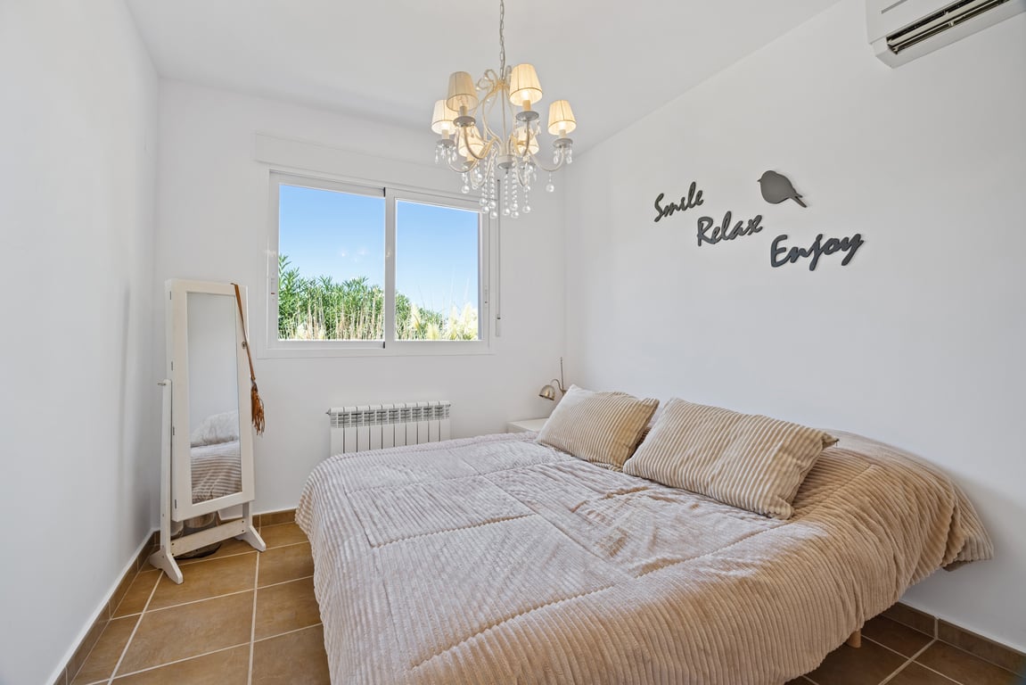 Bright bedroom featuring terracotta tile flooring, split air conditioning, and a radiator. Includes a double-glazed window with garden views and a classic crystal chandelier.