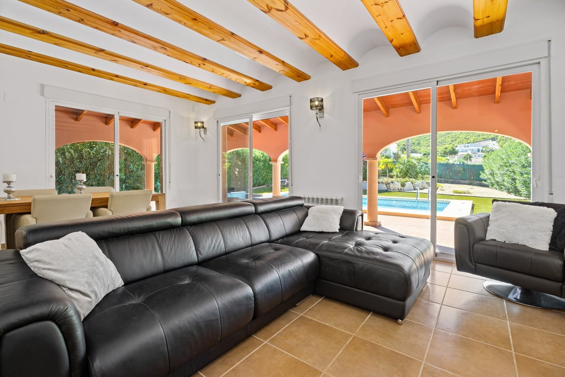 Bright living area with exposed wooden ceiling beams, black leather furniture, and sliding glass doors leading to the pool and mountain scenery.