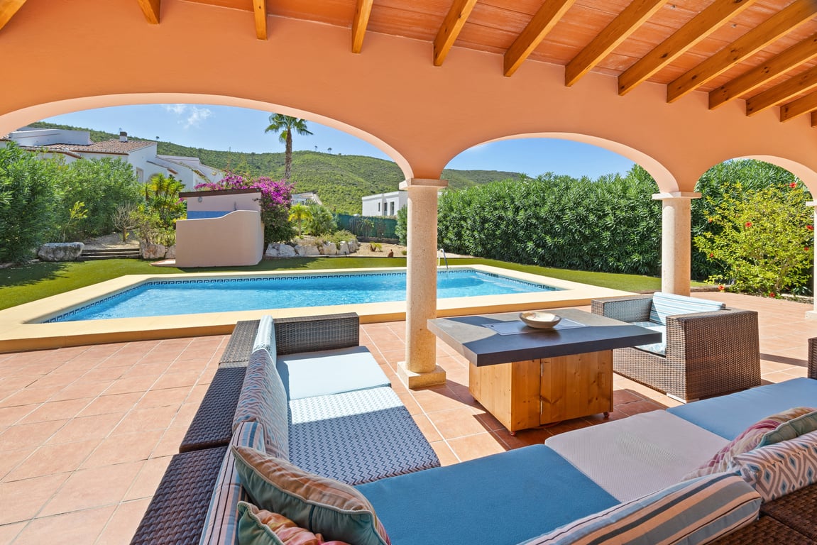 Covered naya featuring wooden beams and stone arches, overlooking a mosaic-tiled pool, artificial lawn, and scenic mountain views.