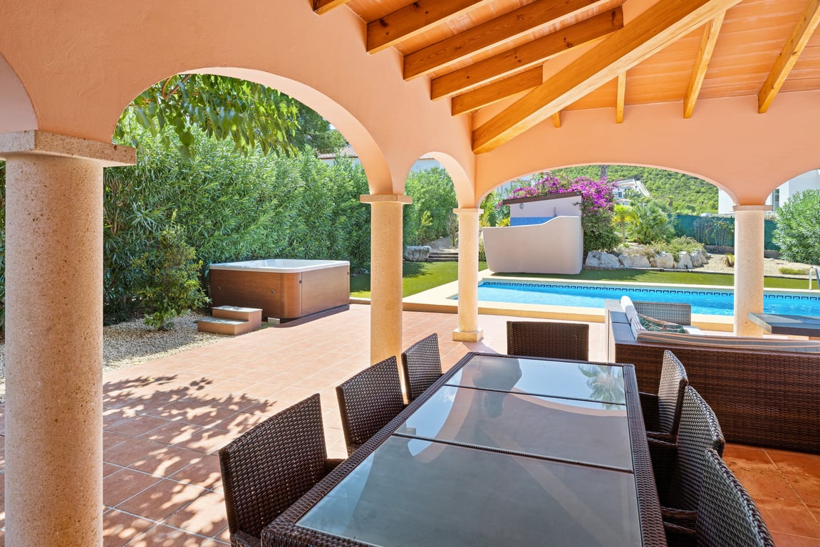 Covered naya featuring wooden ceiling beams and stone pillars, overlooking the mosaic pool, outdoor jacuzzi, and landscaped garden with mountain views.