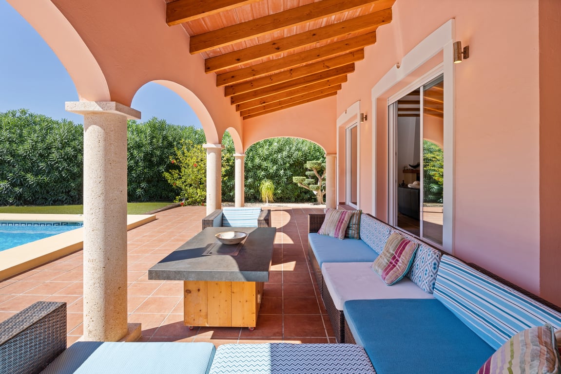 Covered terrace featuring exposed wooden beams, natural stone columns, and a lounge area overlooking the swimming pool and garden.