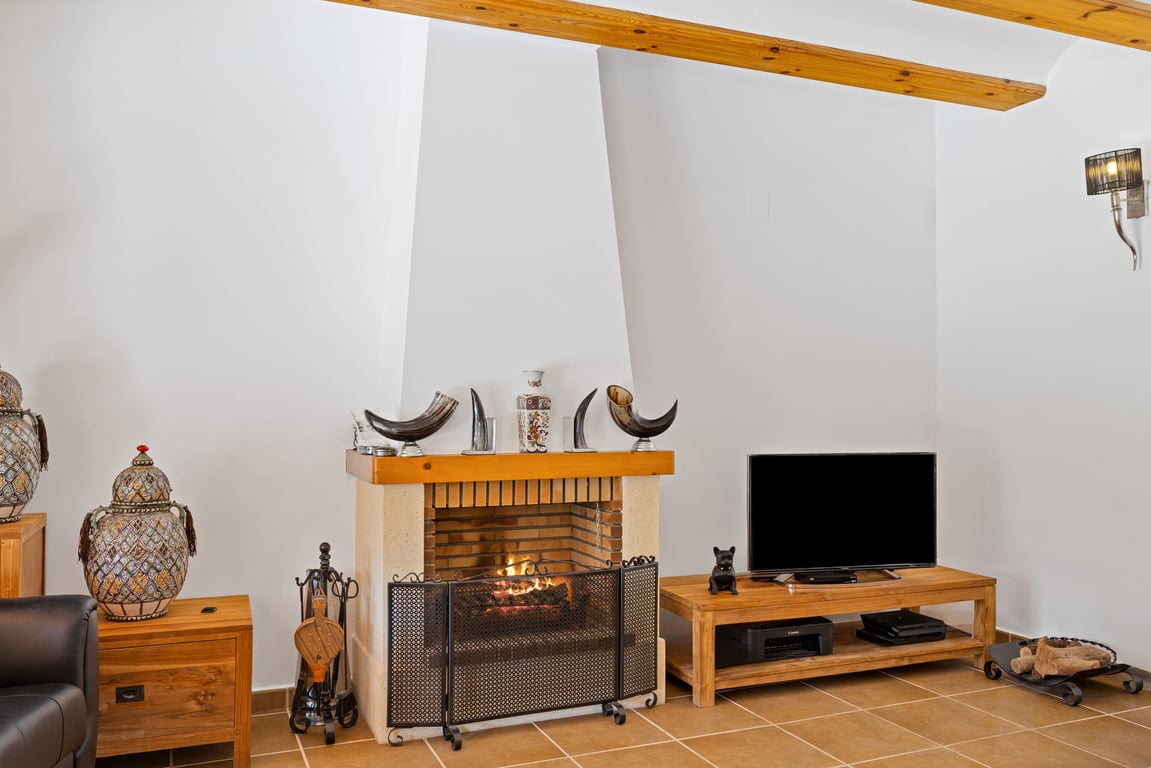 Living area featuring a traditional fireplace with a wooden mantel, exposed ceiling beams, and rustic tiled flooring. A wooden TV stand complements the interior.