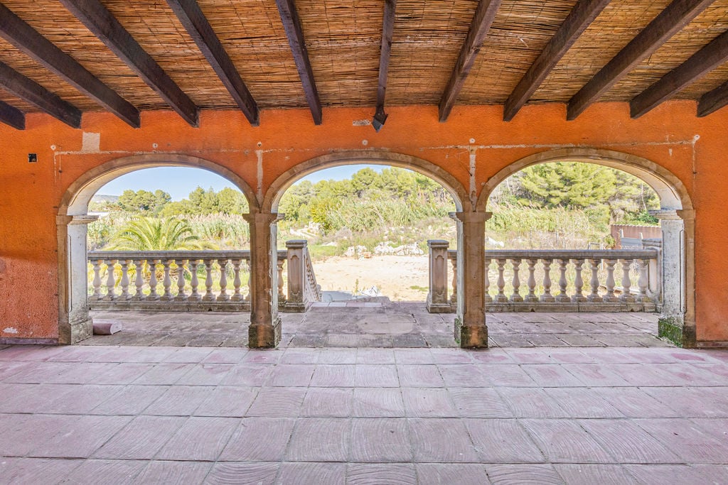 Covered terrace featuring traditional stone arches, exposed wooden beams with cane ceiling, and a classic balustrade overlooking the Mediterranean landscape.