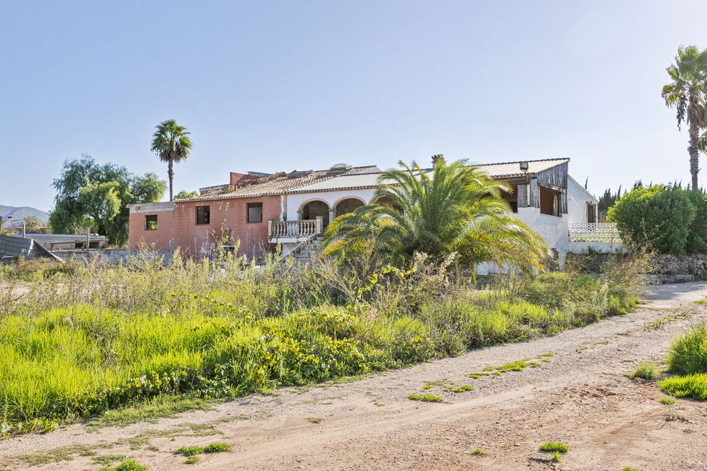 Exterior view of the main structure featuring traditional arches and stone balustrades, set within an expansive rustic plot with mature palm trees and natural greenery.