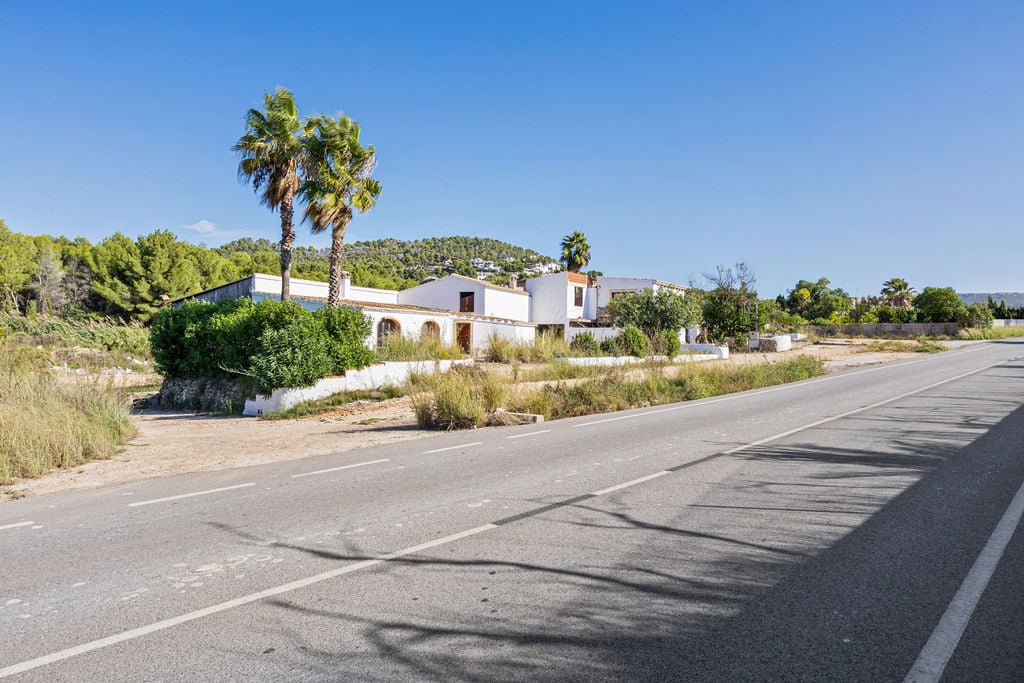 Exterior view of the Mediterranean property from the road access, featuring white facades, palm trees, and a backdrop of wooded hills under a clear sky.