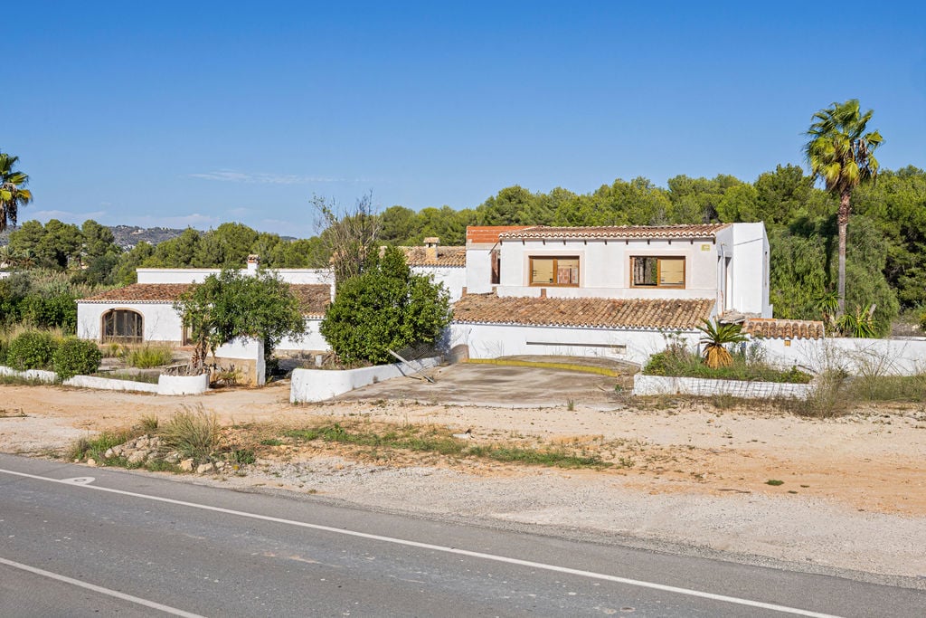 Exterior view of the traditional Mediterranean structure featuring white-washed walls and terracotta roofs, set on a vast plot adjacent to the road.