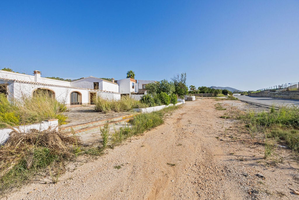 Exterior view of traditional white buildings with arched doorways and tiled roofs, featuring a large unpaved plot with direct roadside access.