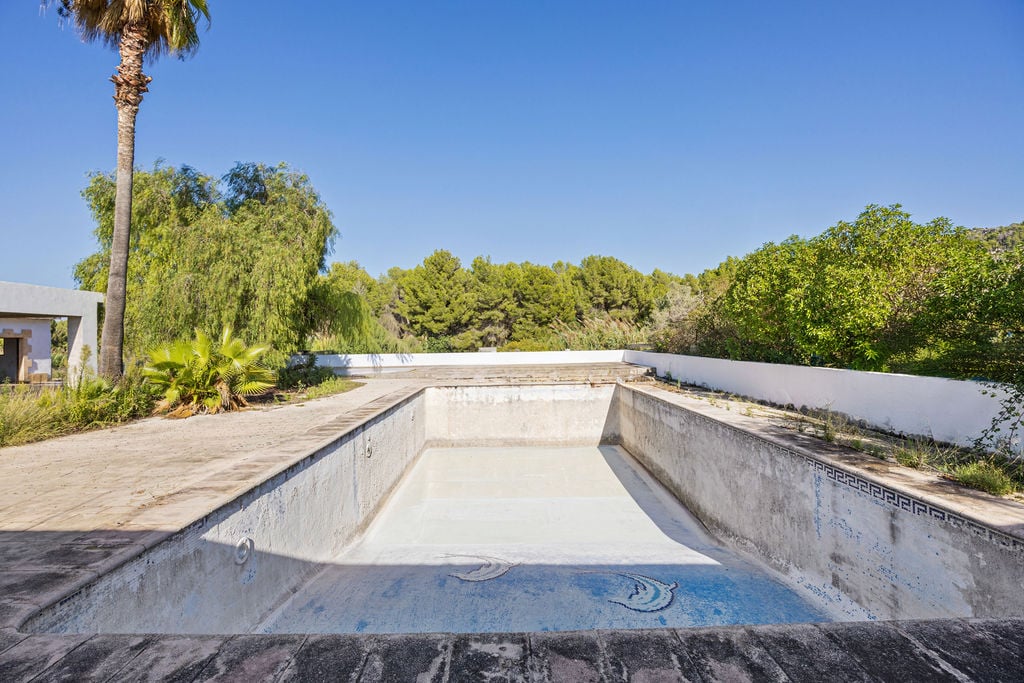 Large rectangular swimming pool awaiting renovation, featuring mosaic detailing and a spacious stone terrace bordered by mature Mediterranean trees.