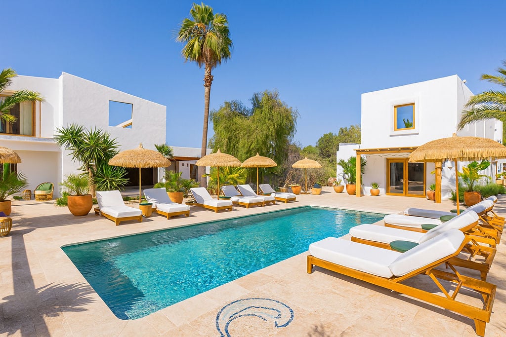Outdoor swimming pool area featuring stone tiling, wooden sun loungers with white upholstery, and heather parasols set against white Mediterranean architecture.