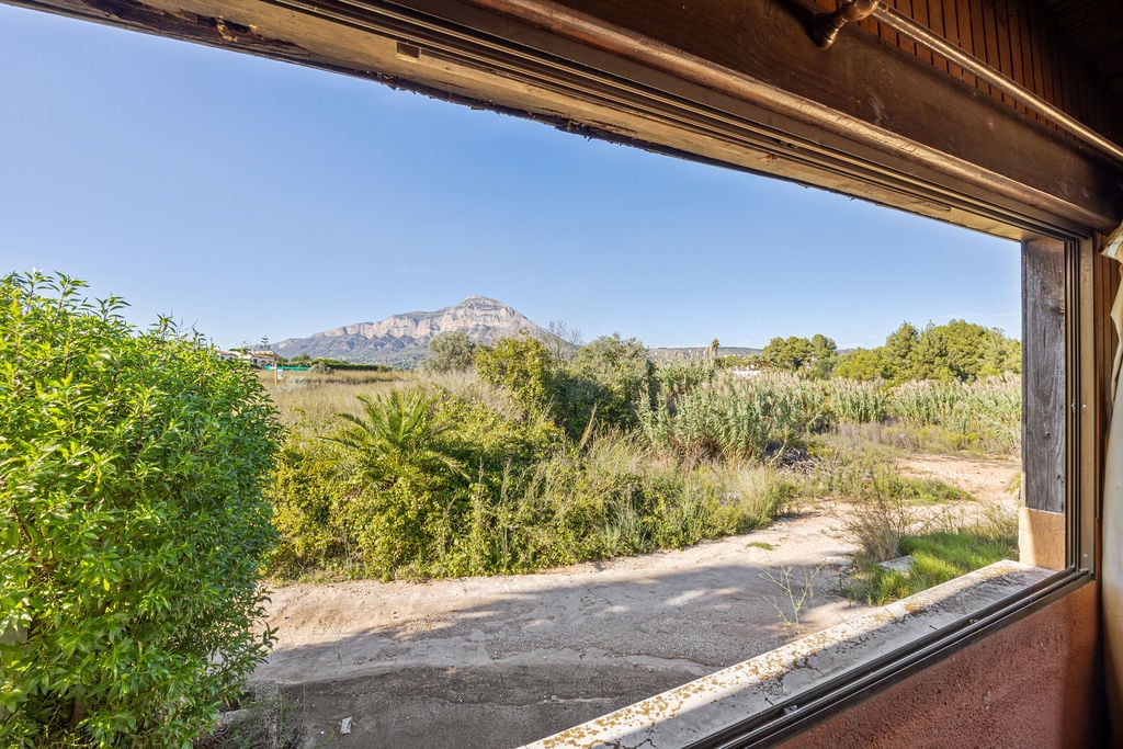 Panoramic view of the Montgó mountain through a wooden-framed window. The foreground features native Mediterranean vegetation and a clear, expansive horizon.