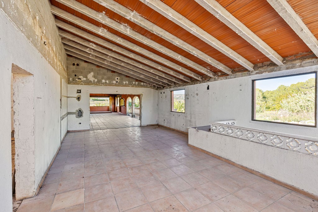 Spacious open-plan interior featuring high vaulted ceilings with exposed concrete beams, terracotta tiles, and large windows overlooking the Mediterranean landscape.