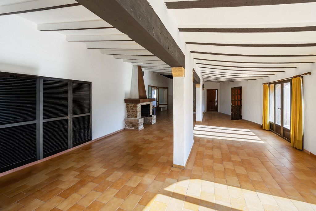 Spacious open-plan room featuring exposed dark wood ceiling beams, terracotta tile flooring, and a traditional stone fireplace. Large windows provide abundant natural light.