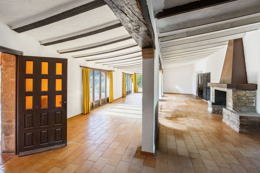 Spacious rustic hall featuring exposed dark wood ceiling beams, traditional terracotta tiling, and a natural stone fireplace with a metal hood.