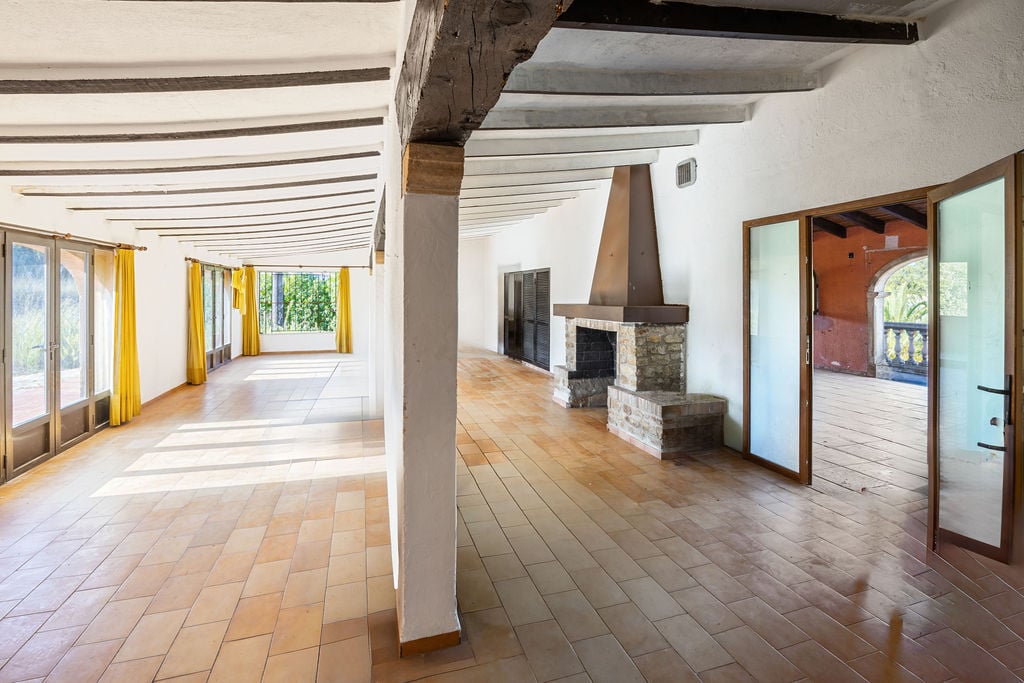 Spacious rustic lounge featuring terracotta tile flooring, exposed wooden ceiling beams, and a stone fireplace. Large windows and glass doors provide abundant natural light.