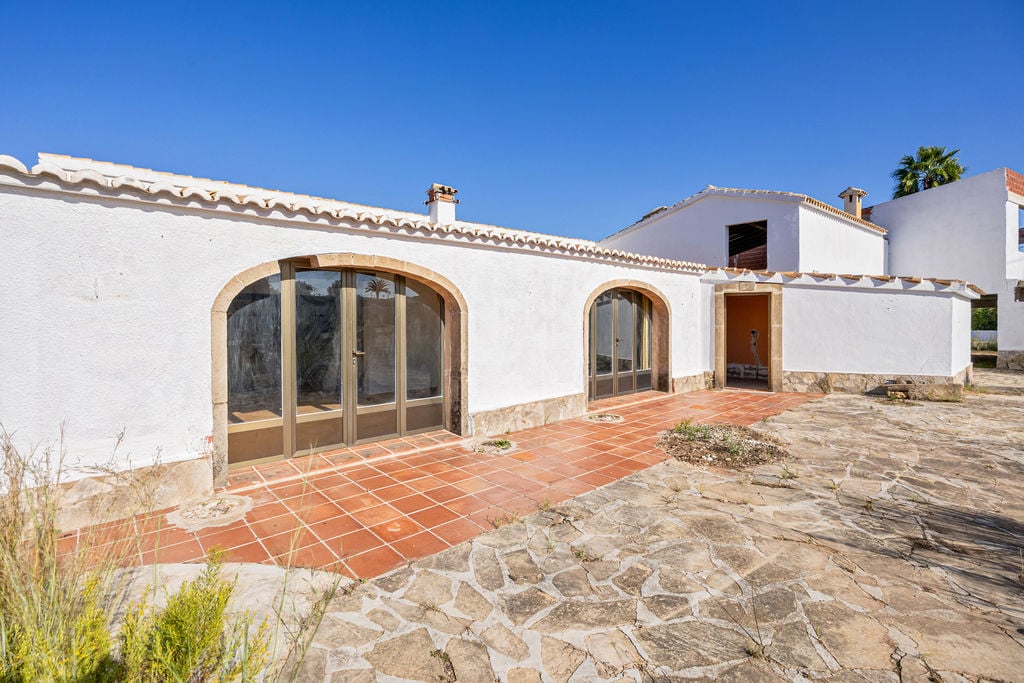 White facade featuring traditional stone-framed arches and large glass doors. The exterior transitions from terracotta tiles to rustic natural stone paving.