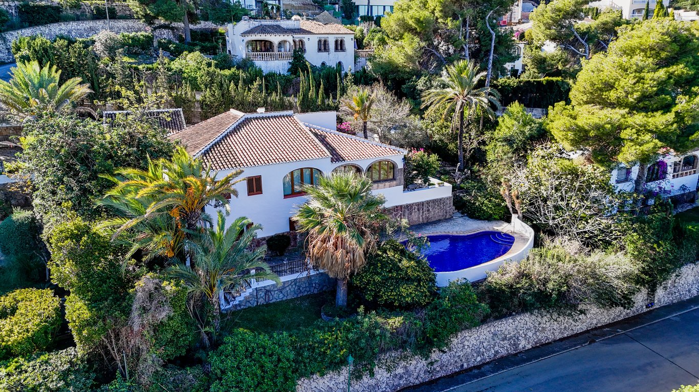 Aerial view of the traditional villa featuring a kidney-shaped pool, arched naya, and lush Mediterranean gardens enclosed by natural stone walls.