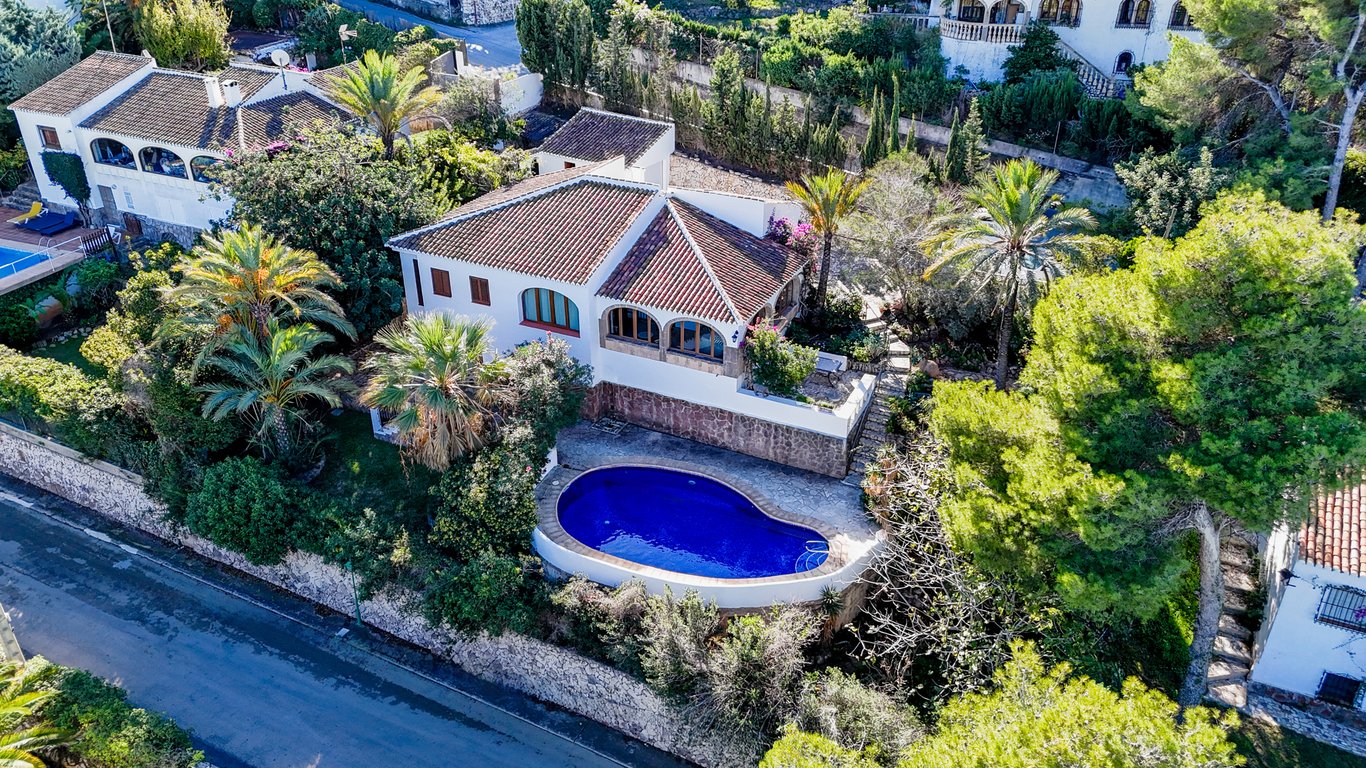 Aerial view of the traditional villa featuring a kidney-shaped pool, terracotta roof, and arched naya, set within a mature Mediterranean garden with palm trees.
