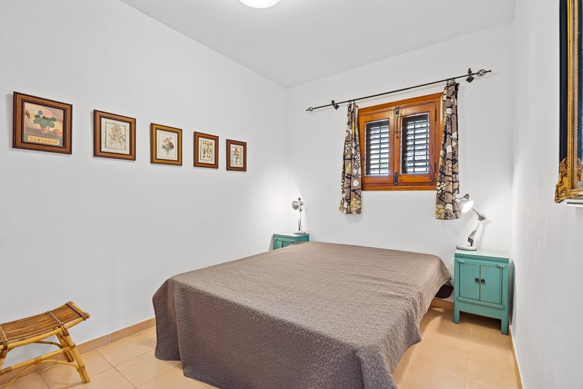 Bedroom featuring a double bed, turquoise bedside tables, and a wooden window with traditional shutters. White walls are accented by a series of botanical prints.