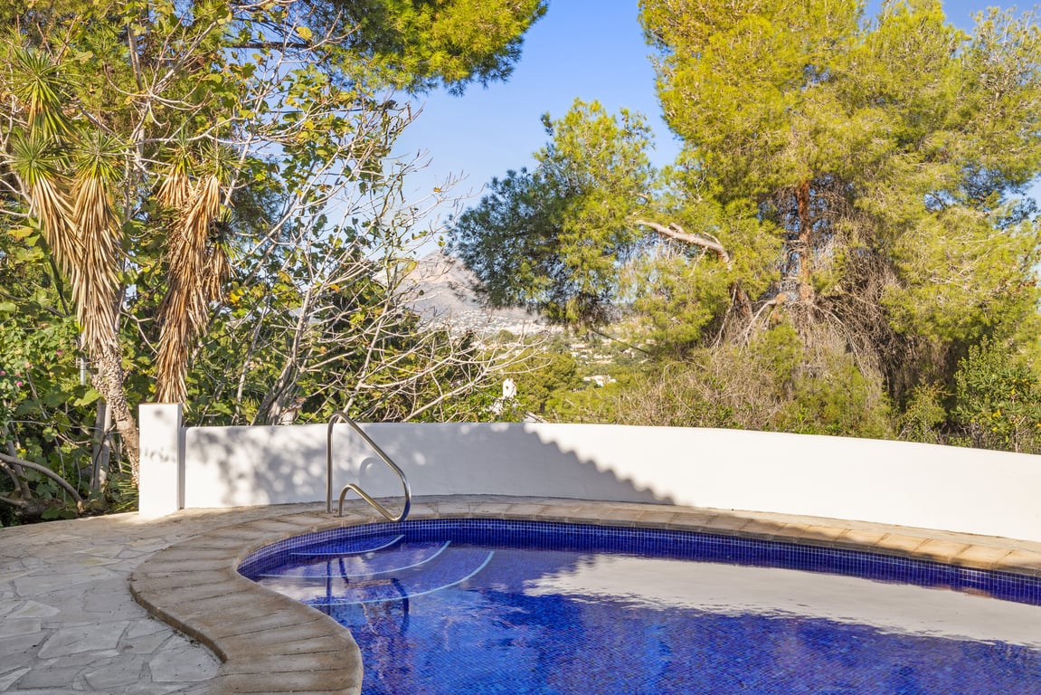 Blue tiled swimming pool with metal handrail, surrounded by a natural stone terrace and lush Mediterranean trees with distant views of the Montgó mountain.
