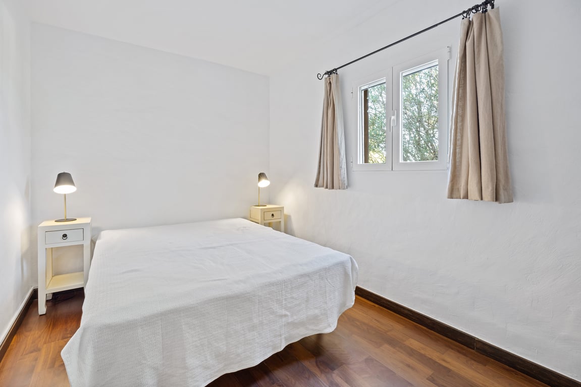 Bright bedroom featuring wooden flooring, white textured walls, and a double-glazed window overlooking greenery. Includes bedside tables and reading lamps.