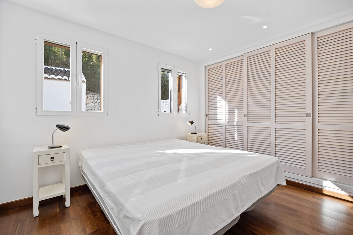 Bright bedroom featuring wooden flooring, white walls, and a large built-in wardrobe with slatted sliding doors. Equipped with double-glazed windows and recessed ceiling lighting.