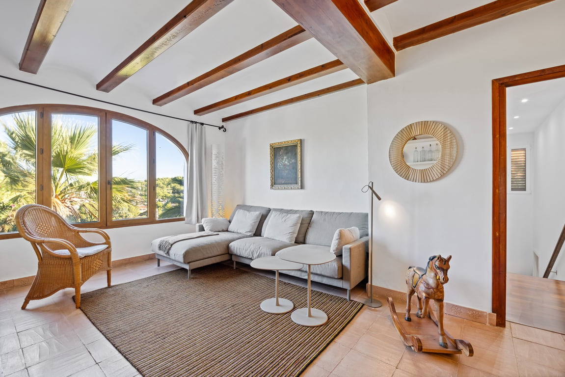 Bright living area featuring exposed wooden ceiling beams, traditional ceramic flooring, and an arched window overlooking Mediterranean greenery.