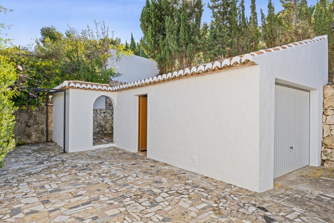 Detached garage and utility building featuring traditional white plaster, terracotta roof tiles, and irregular stone mosaic paving under mature cypress trees.
