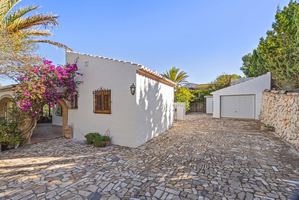 Exterior entrance featuring an irregular stone-paved driveway, detached garage, and white facade adorned with pink bougainvillea and traditional Tosca stone arches.